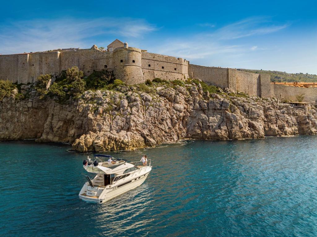 Jachta peld pa skaidrām ūdeņiem pie klints, kur redzama senatnīga cietokšņa mūra konstrukcija, kas ieskauj krastu.