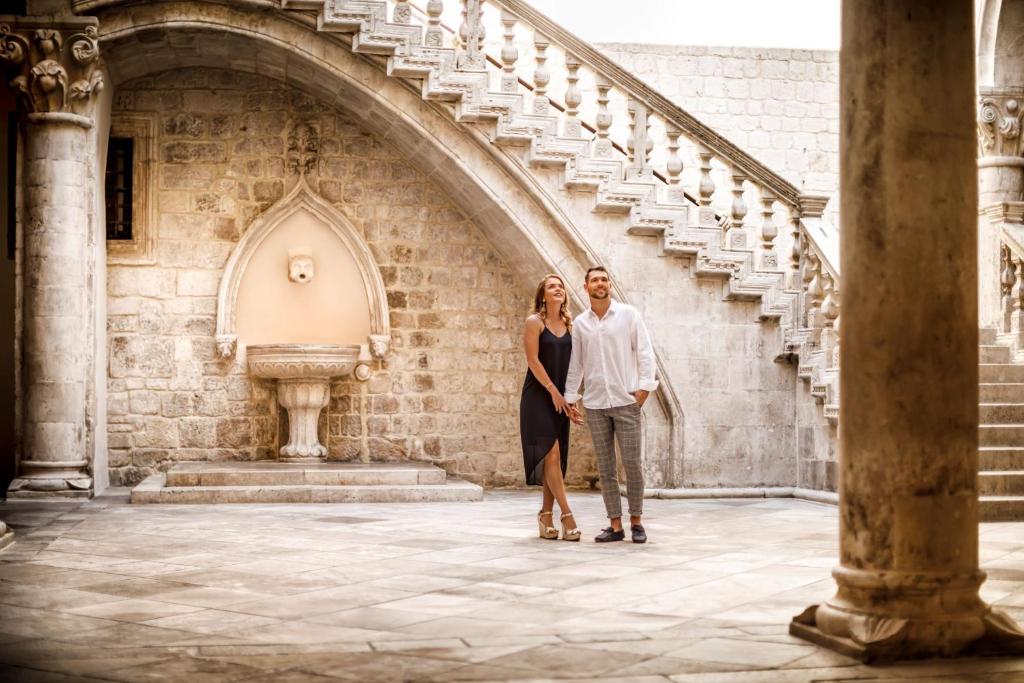 Romantisks pāris stāv gleznainā iekšpagalmā ar akmens sienām un greznām kāpnēm, baudot kopīgu brīdi. Fons ietver dekoratīvu strūklaku un arhitektoniskus elementus.