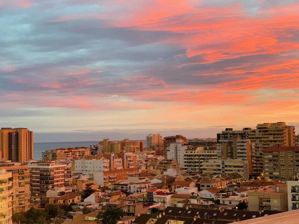 Pilsētas ainava ar krāšņu saulrietu, kur debesīs redzami rozā un violeti toņi. Skats uz ēkām un jūru, kas piešķir atmosfēru.