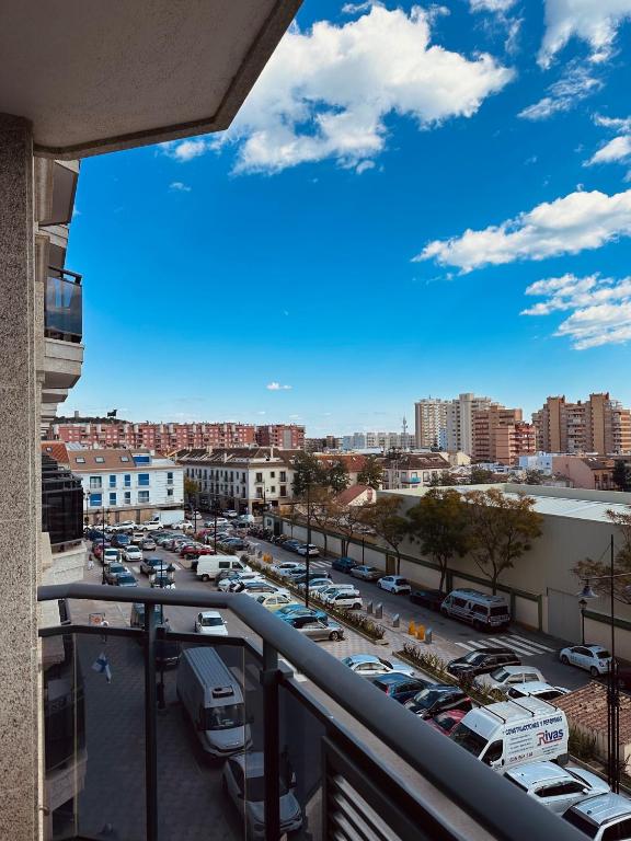 Skats no balkona uz pilsētas ielu, kur redzamas automašīnas un dzīvojamās ēkas. Debesīs plūstoši mākoņi un saulains laiks.
