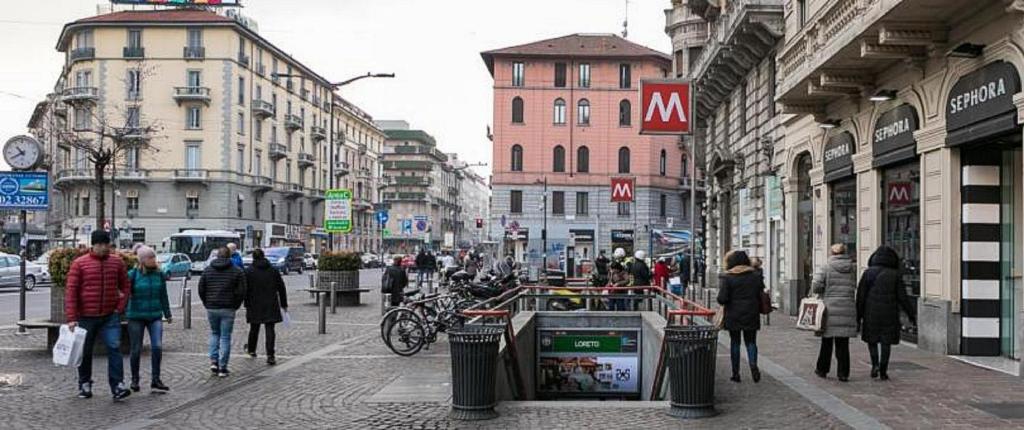 Pilsētas iela, kurā redzami gājēji, veikali un metro ieeja. Apkārtne ir rosīga, ar velosipēdiem un transportlīdzekļiem.