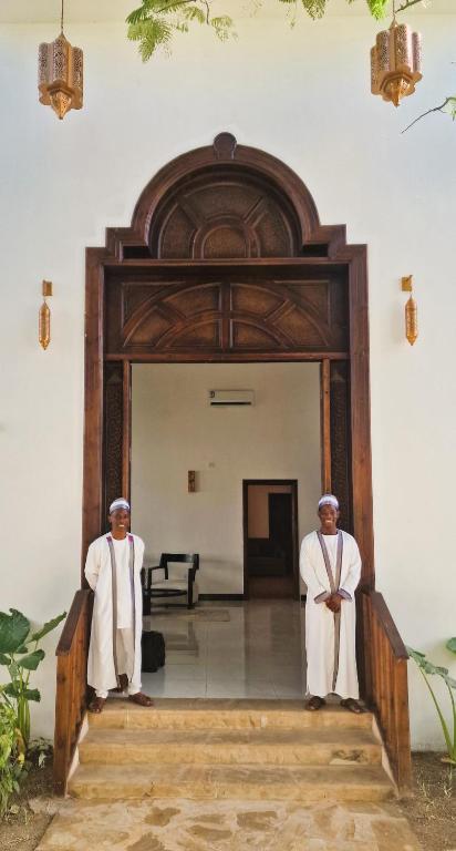 Divi vīrieši tradicionālās drēbēs stāv pie ieejas elegantā mājā ar greznu koka durvju rāmjiem. Apkārt ir zaļumi un dekoratīvi gaismas ķermeņi.