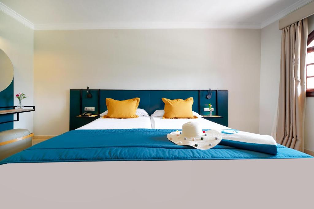 Mūsdienīga viesnīcas istaba ar divām gultām, spilveniem un zilu gultas pārklāju. Uz gultas atrodas vasaras cepure un dvielis, kā arī neliels zieds uz galda.