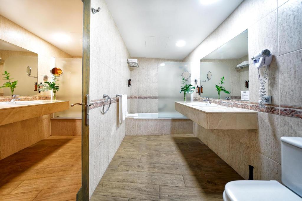 Moderns un elegants vannas istabas interjers ar divām izlietnēm un vannu, kas apgaismota ar siltu gaismu un zaļām augu kompozīcijām, radot relaksējošu atmosfēru.