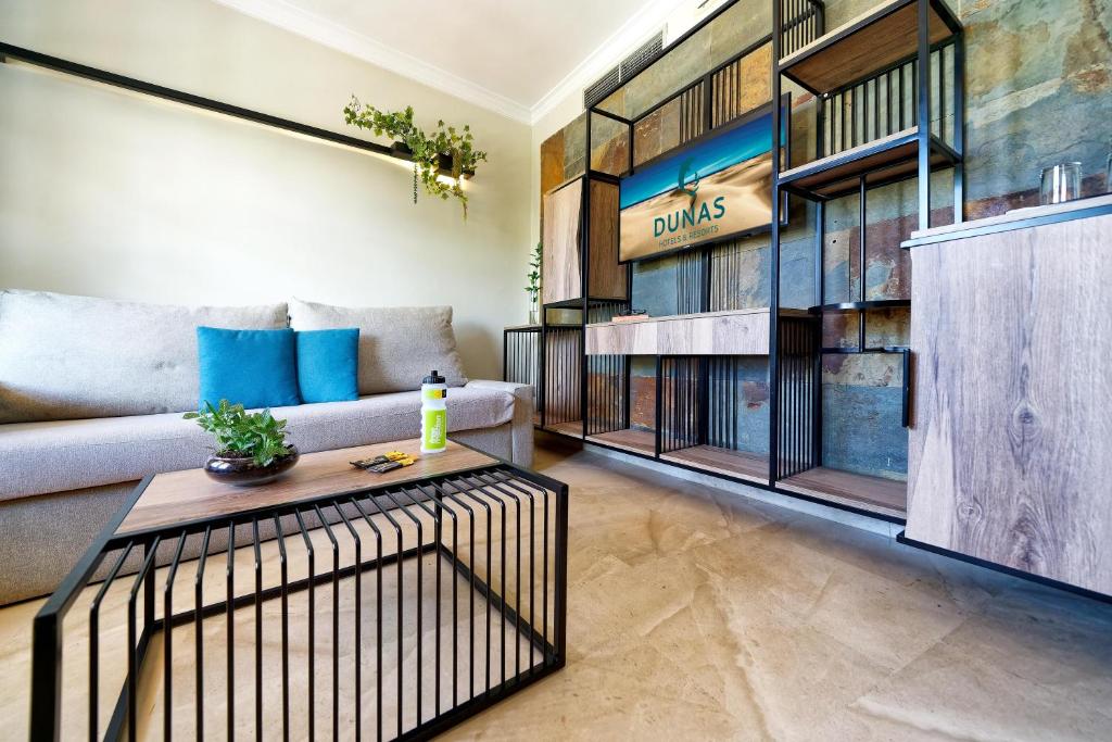 Mūsdienīgs viesistabas interjers ar pelēku dīvānu, zilu spilvenu un koka galdiņu, kas rotāts ar augiem. Sienas dekorētas ar stilīgu plauktu sistēmu un televizoru, uz kura redzams zīmols "DUNAS".