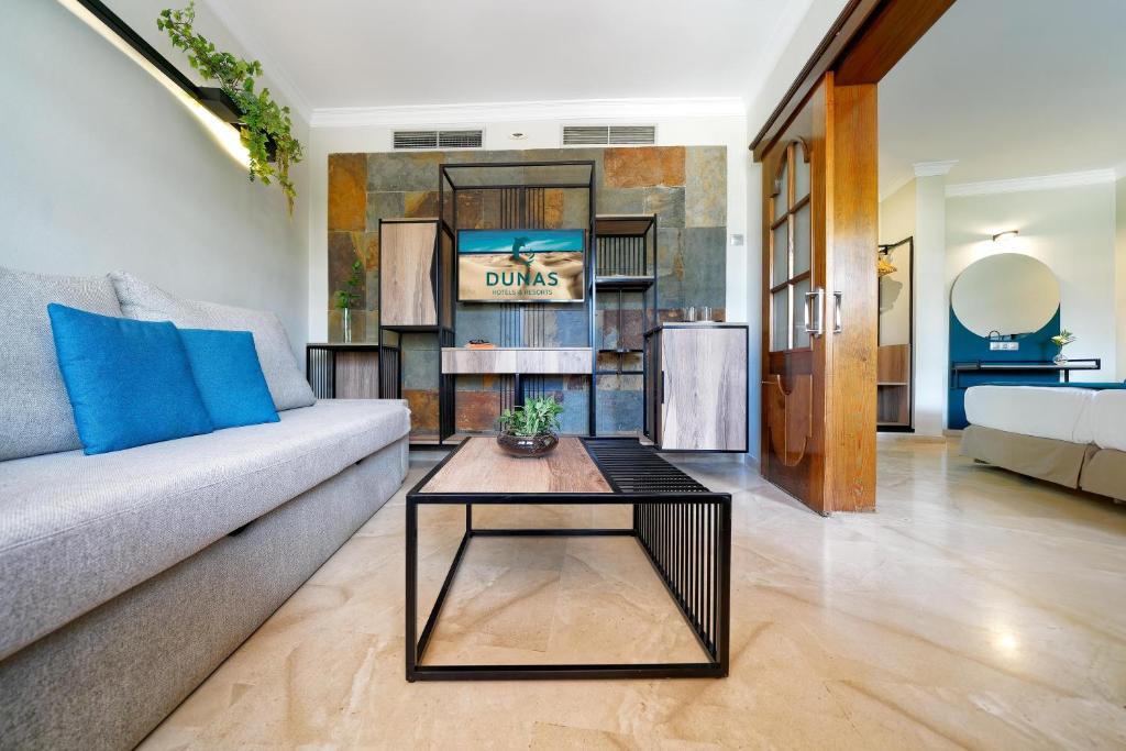 Mūsdienīgs viesnīcas numurs ar elegantu mēbeļu dizainu, pelēku dīvānu un koka kafijas galdiņu, kas papildina interjeru. Uz sienas redzama viesnīcas "Dunas" zīme.