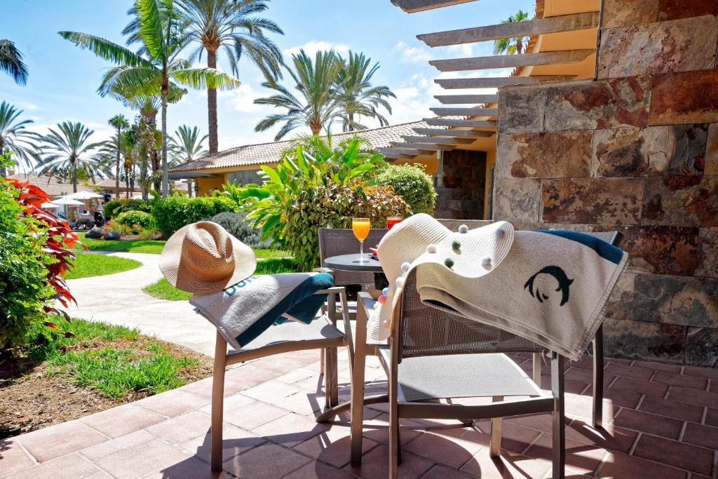 Atpūtas stūris ar krēsliem, uz kuriem atrodas cepure un dvielis, apkārt zaļojoši palmu koki un krāsaini ziedi, ideāla vieta relaksācijai pie baseina. Uz galda stāv dzēriens, radot brīvdienu atmosfēru.