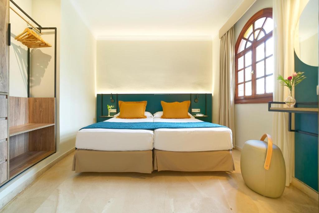 Mūsdienīgs guļamistabas interjers ar divām gultām, dekorēts ar zeltainām spilveniem un zilām gultas pārklājiem. Telpā ir liels logs, kas nodrošina dabiski apgaismojumu, kā arī elegantas mēbeles un dekoru.