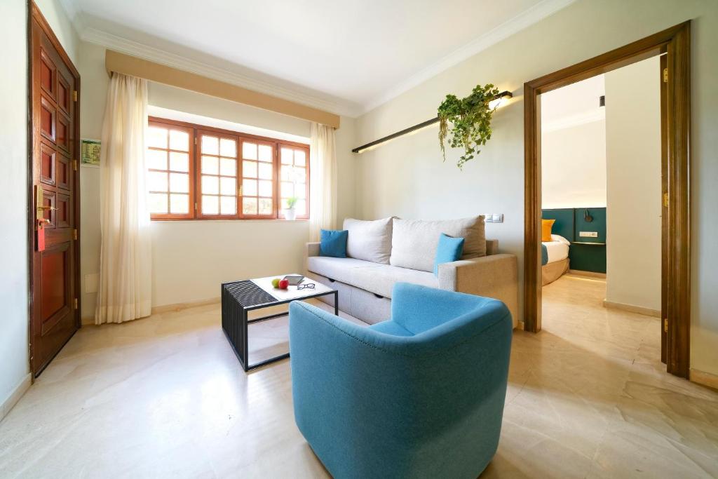 Moderns viesistabas interjers ar ērtu dīvānu, krēslu un lieliem logiem, kas nodrošina dabisko apgaismojumu. Telpā ir arī dekoratīvas augu kompozīcijas un piekļuve guļamistabai.