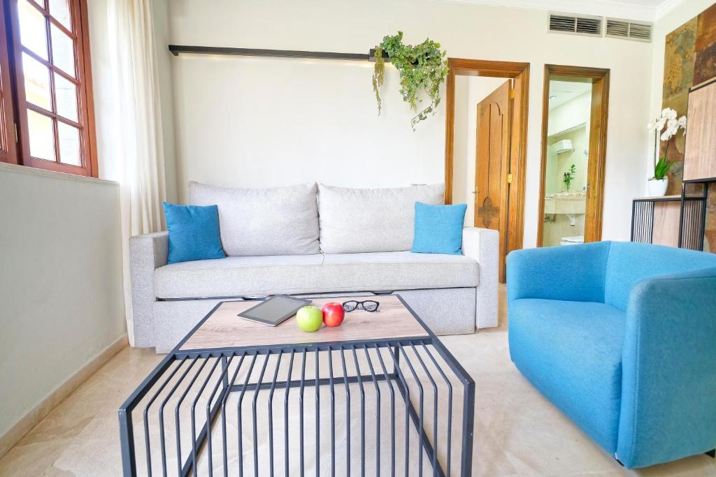 Mūsdienīga viesistaba ar pelēku dīvānu, zilu krēslu un melnu metāla galdu, uz kura atrodas planšetdators un augļi. Interjerā ir gaišas krāsas un dabiski elementi, radot patīkamu un ērtu atmosfēru.