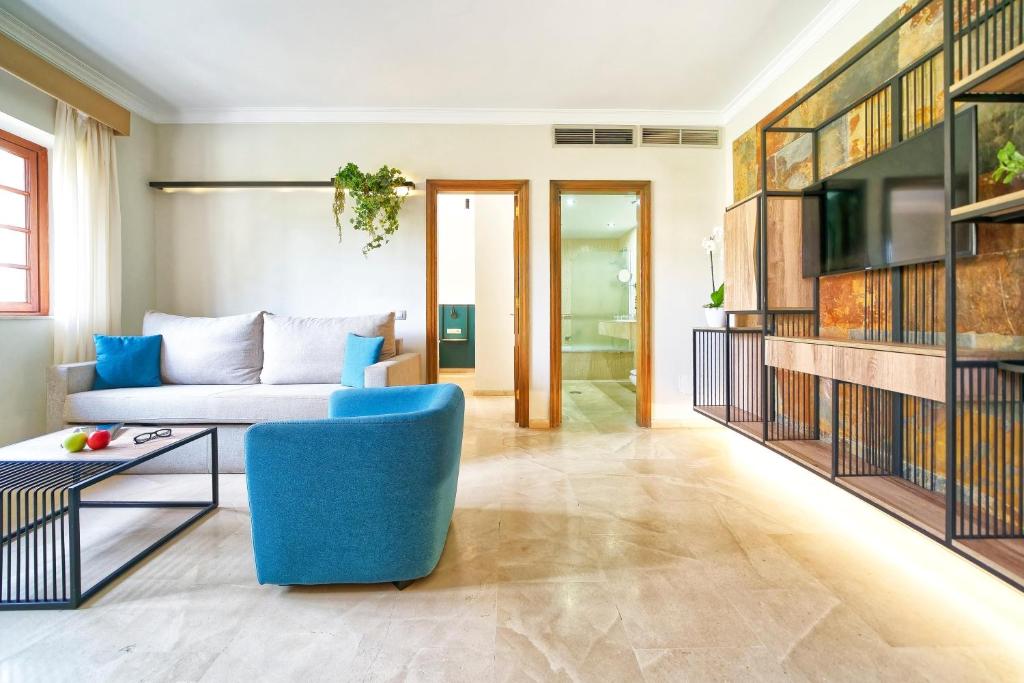 Mūsdienīgs viesistabas interjers ar ērtu dīvānu, spilveniem un zilu krēslu, dekorēts ar augu elementiem un mūsdienīgu TV stendu. Gaisma un siltas krāsas rada mājīgu atmosfēru.