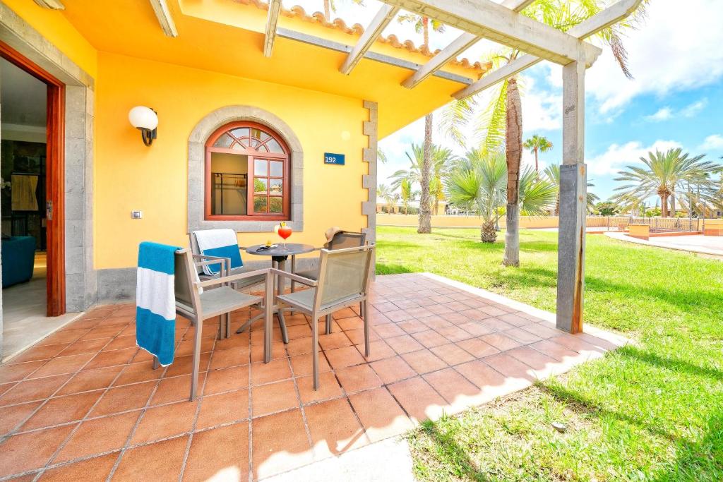 Mūsdienīga terase ar galdu un krēsliem, kurā redzams dzēriena glāze un pludmales dvielis, apkārt zaļojoša zāle un palmas. Ideāla vieta relaksācijai saulainā dienā.