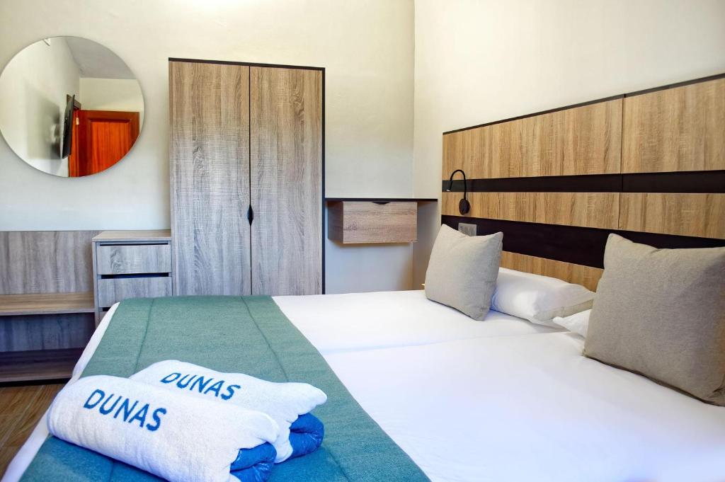 Mūsdienīga viesnīcas numura iekārtojums ar divām gultām, koka mēbelēm un apgaismojumu. Uz gultām redzamas dvieļi ar uzrakstu "DUNAS".