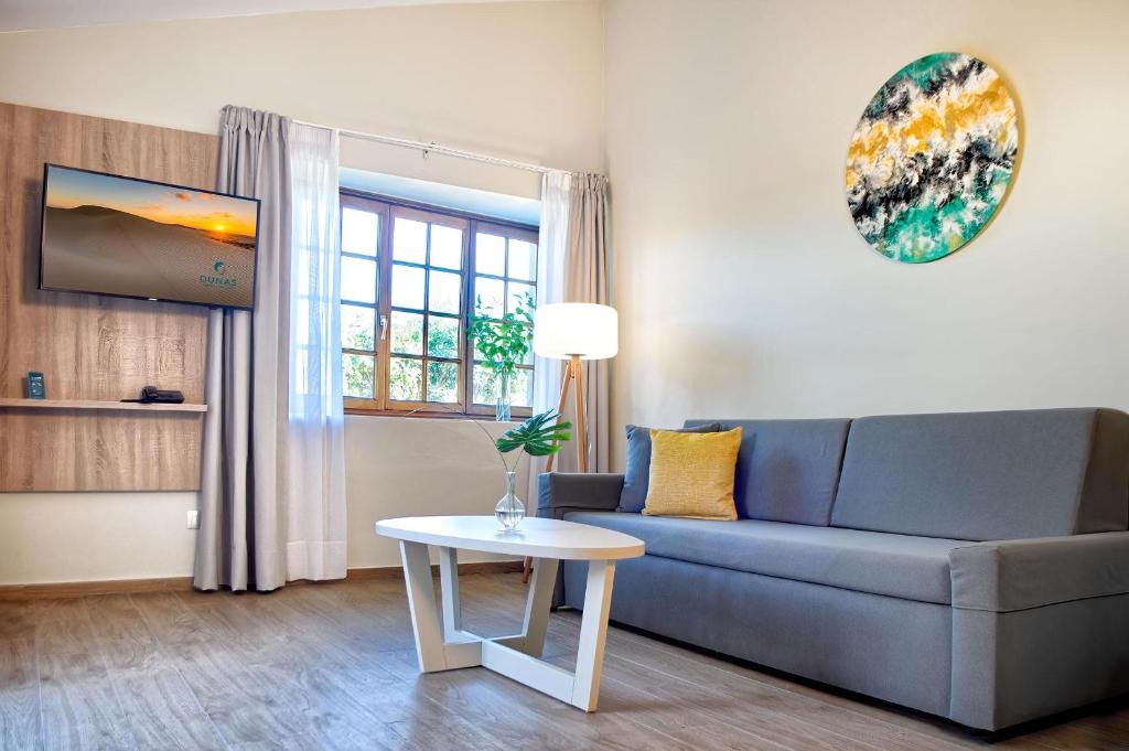 Mūsdienīgs dzīvojamās telpas interjers ar pelēku dīvānu, baltu kafijas galdiņu un televizoru uz koka sienas. Logi ar daudzām rāmēm ļauj ienākt dabiskajai gaismai, radot mājīgu atmosfēru.