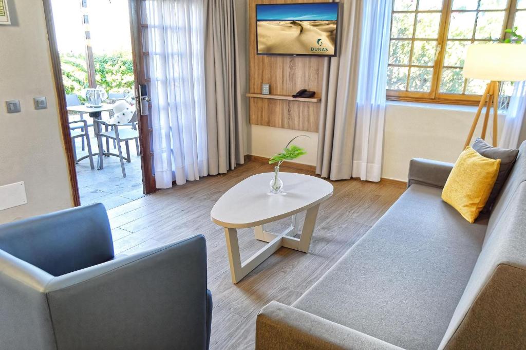 Mūsdienīga viesnīcas istaba ar ērtu dīvānu, krēslu un galdu. Logā redzama terase un dārza ainava, bet uz sienas ir televizors.