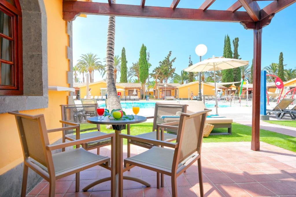 Paviljons ar skatu uz baseinu, kurā redzami krēsli un galds ar dzērieniem un augļiem, apkārt palmām un saulainai atmosfērai.