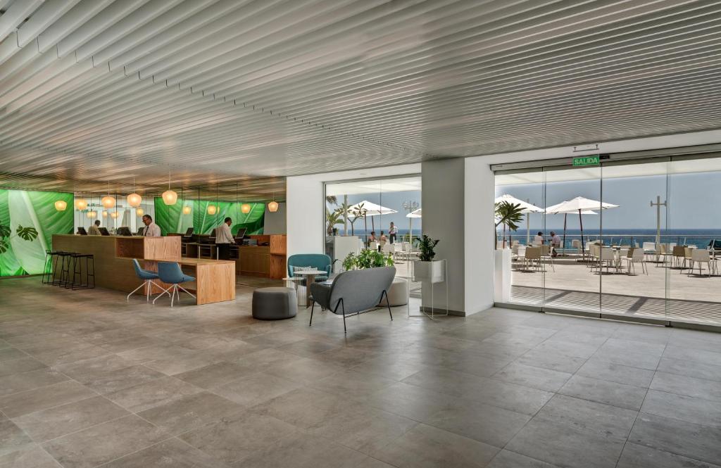 Mūsdienīgs viesnīcas reģistratūras interjers ar zaļām akcentu sienām, modernām mēbelēm un skatu uz jūru caur lieliem logiem. Viesi var baudīt atpūtu terasē zem saulessargiem.