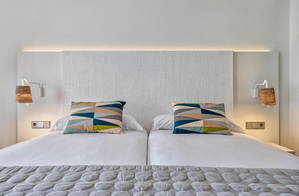 Mūsdienīgs guļamistabas interjers ar divām gultām, dekoratīviem spilveniem un siltu apgaismojumu. Gultas galvgala dizains ir elegants un teksturēts, radot patīkamu atmosfēru.