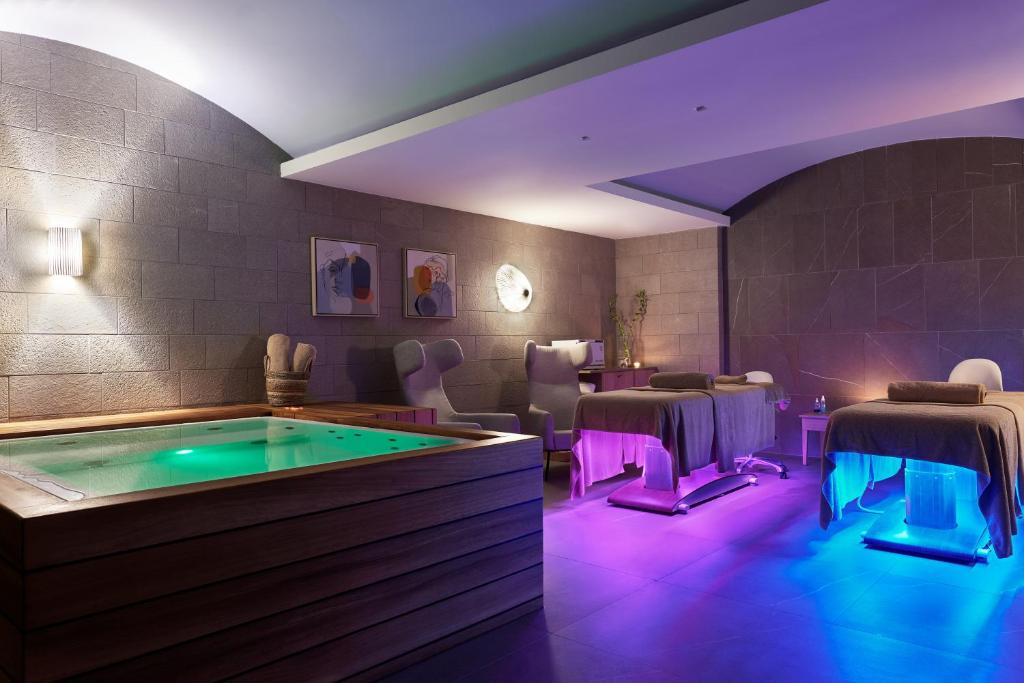 Mūsdienīgs spa telpas interjers ar burbuļvannu un masāžas galdiem, kas apgaismoti ar krāsainām gaismām. Telpa rada relaksējošu un mierīgu atmosfēru.