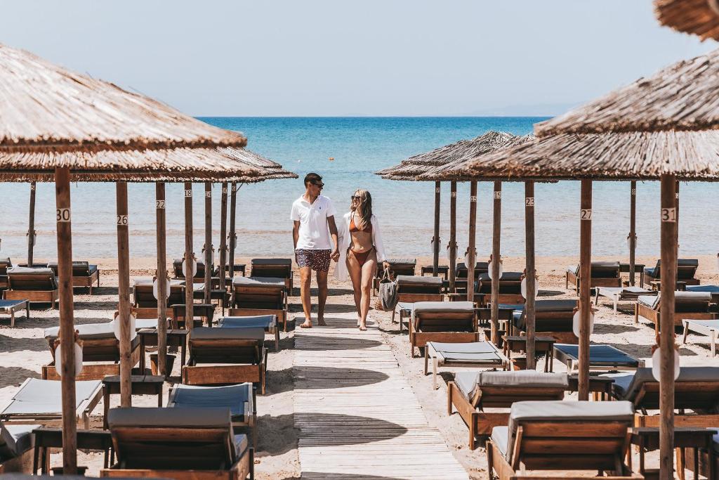 Jauka pāra pastaiga pa pludmali, gar šikiem sauļošanās krēsliem un saulessargiem, fona zilais jūras ūdens un skaidrs debesis.