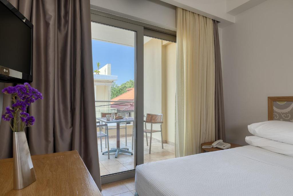 Mūsdienīgs viesnīcas numurs ar balkonu, televizoru un gultu, papildināts ar ziedu pušķi uz koka galda. 