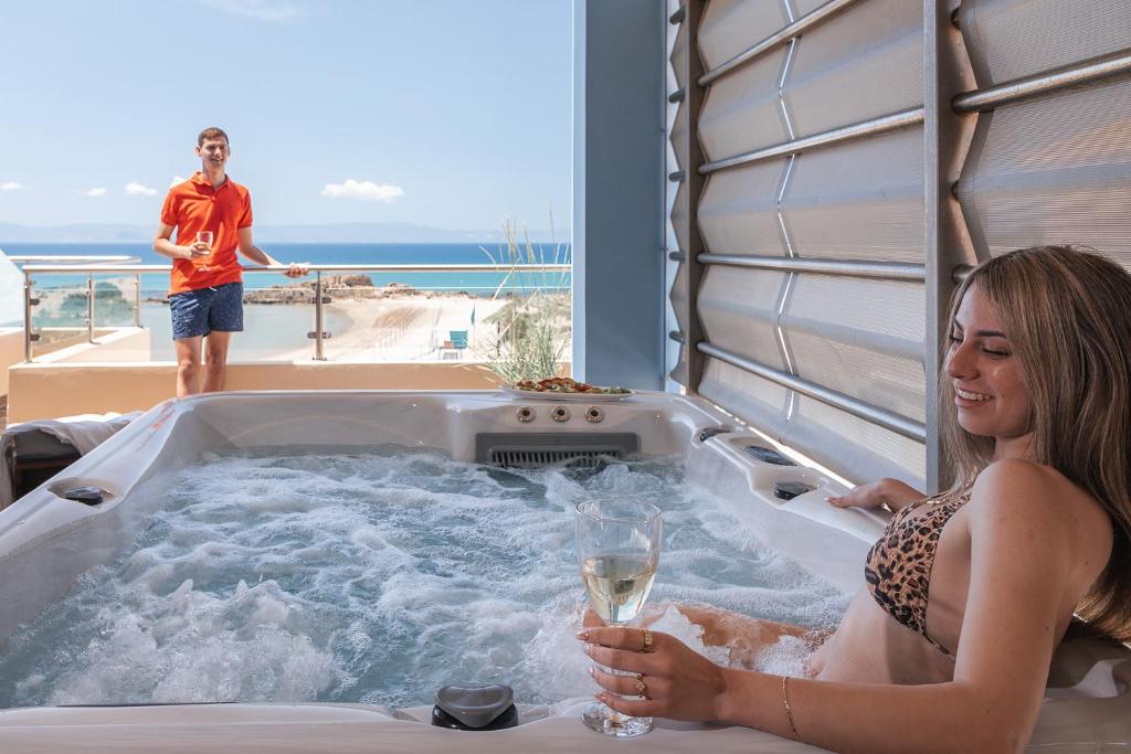 Relaksējoša brīvdienu aina ar sievieti, kas bauda burbuļvannu, un vīrieti, kurš stāv blakus ar dzērienu rokā, fons ir jūras skats un saulains laiks.