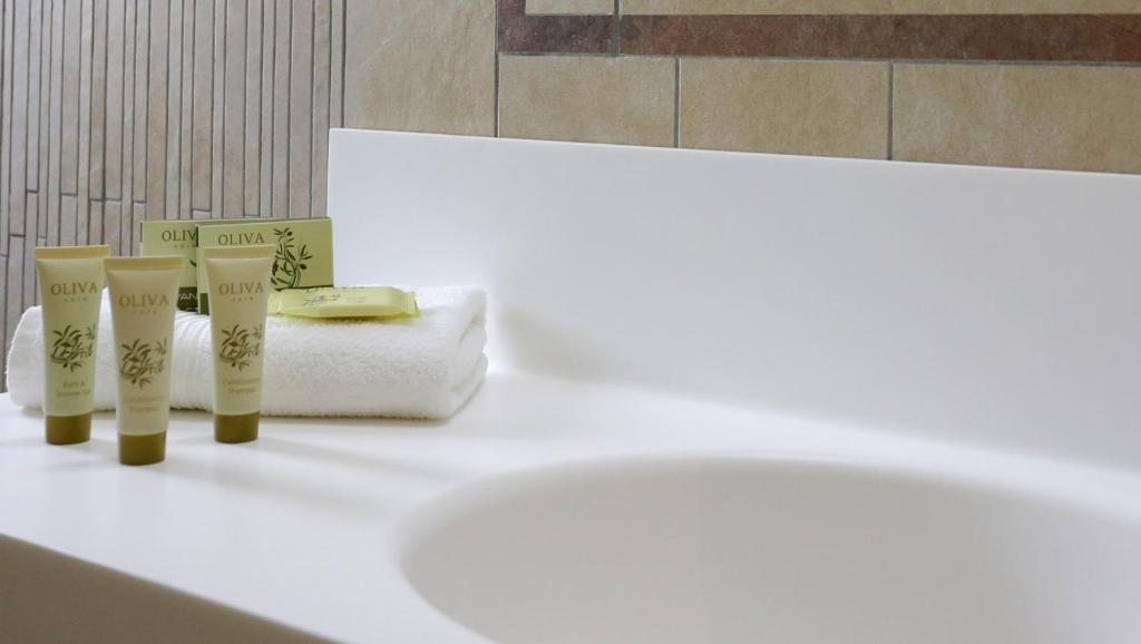 Baltā vannas istabas virsma ar OLIVA zīmola kosmētikas produktiem un dvieli, radot relaksējošu un elegantu atmosfēru.