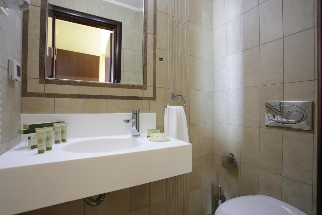 Mūsdienīgs un elegants vannas istabas interjers ar gaišām flīzēm, baltu izlietni un dabīgiem higiēnas produktiem uz plaukta. Spogulis un mīksta dvieļu turētājs papildina telpas estētiku.