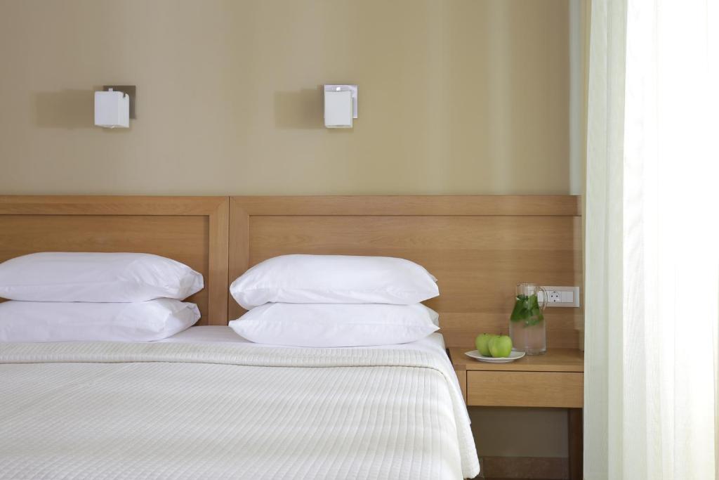 Mūsdienīga guļamistaba ar koka gultu un baltiem spilveniem, blakus atrodas naktsgaldiņš ar ūdens karafēm un zaļām ābolu šķēlītēm.