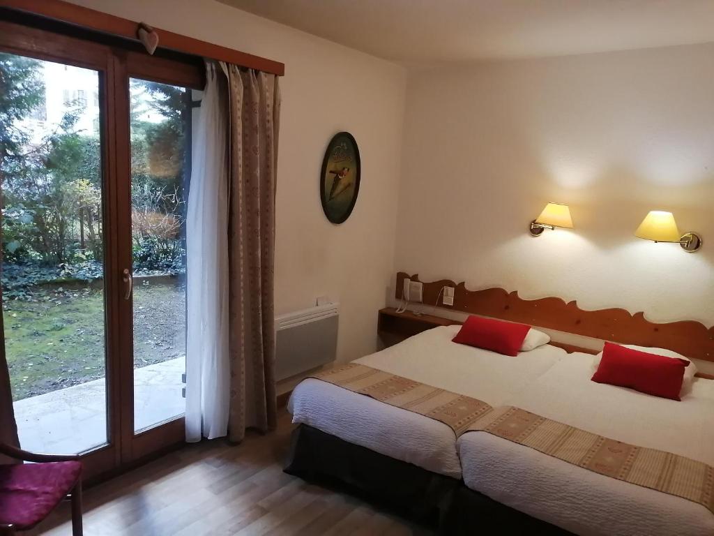 Gaisma un ērta viesnīcas istaba ar divām atsevišķām gultām, sarkaniem spilveniem un skatu uz dārzu caur lieliem logiem. Interjers ir vienkāršs, ar siltām krāsām un koka elementiem.