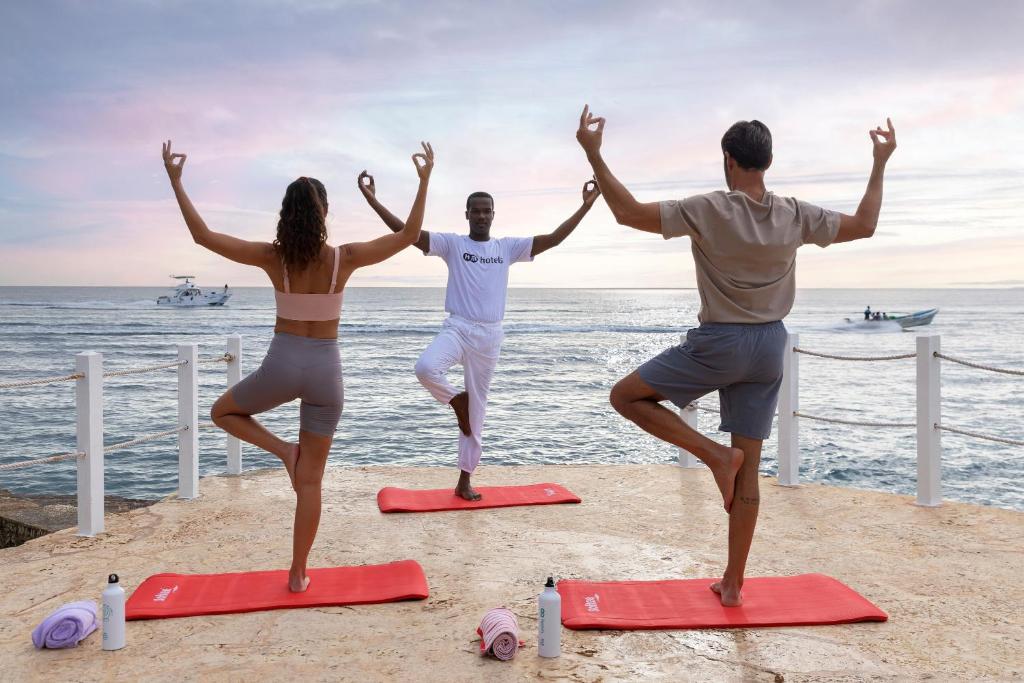 Joga nodarbība pie jūras saulrietā, kur divi cilvēki seko instrukcijām no pasniedzēja. Visi trīs ir uz jogas paklājiem un veic līdzsvara pozas.