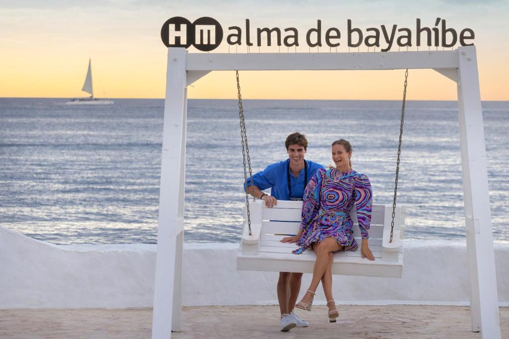 Pāris sēž baltā šūpuļtīklā, ar saulrietu fona un jūras viļņiem aiz muguras. Virs viņiem redzams "Alma de Bayahibe" zīme.