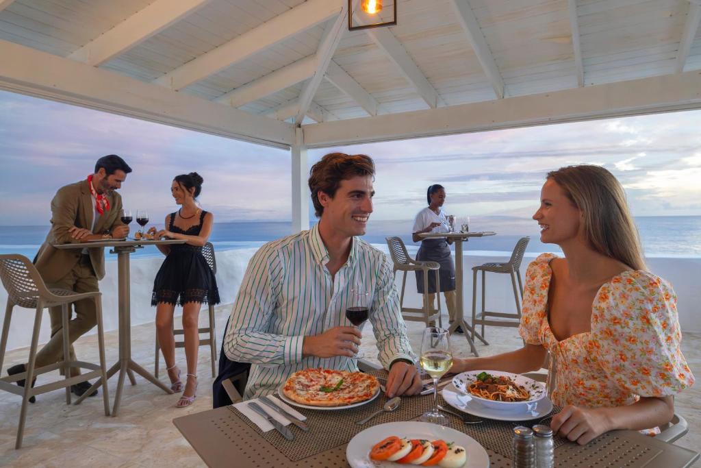 Romantiska vakariņas pie jūras, kur pāris bauda ēdienus un vīnu, kamēr citi viesi izbauda atmosfēru. Gaisotne ir eleganta un relaksējoša, ar skatu uz okeānu.