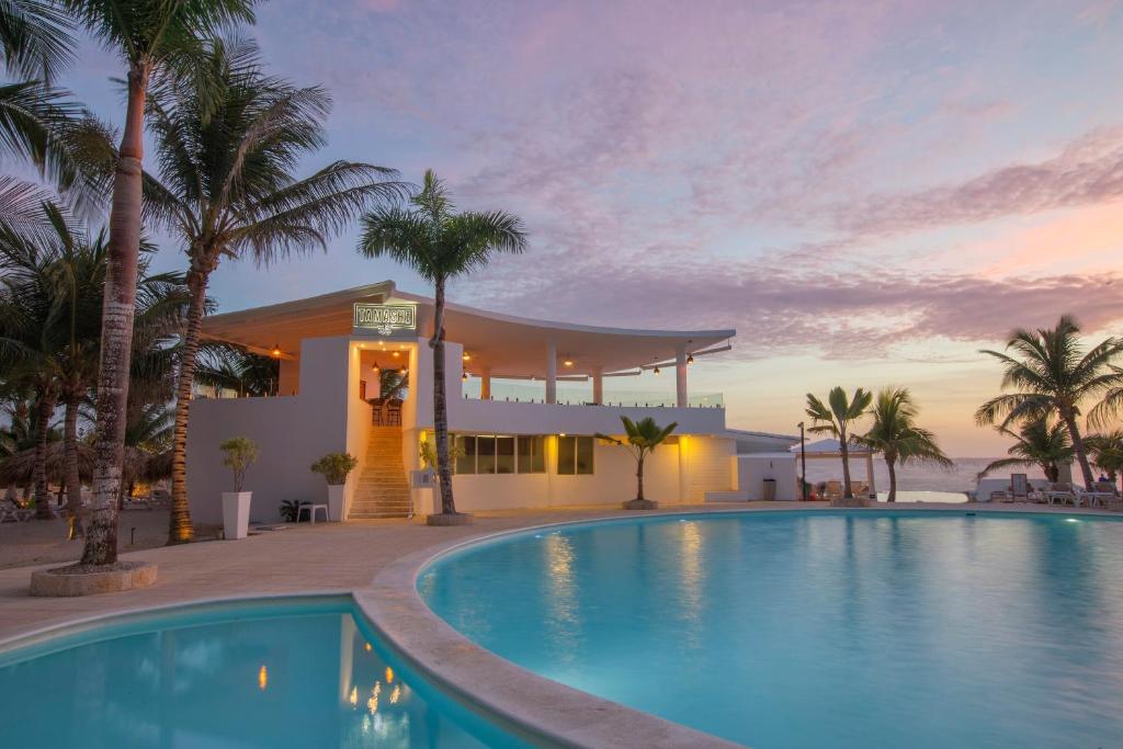 Elegants baseins ar palmām, kas atrodas pie modernā restorāna ar skatu uz saulrietu. Mājas fasāde ir gaiša un mūsdienīga, radot relaksējošu atmosfēru pludmales vidē.