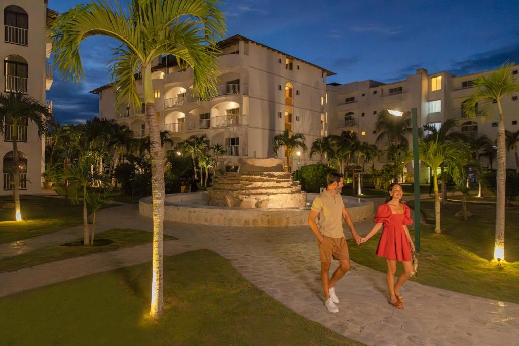 Romantisks vakara brīdis pāri, kas pastaigājas gar strūklaku, apgaismotiem dārziem un modernām ēkām. Siltas gaismas un tropiskie augi veido patīkamu atmosfēru.