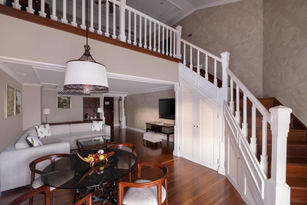 Mūsdienīgs viesistabas interjers ar gaišām krāsām, elegantu galdu un trepēm, kas ved uz otro stāvu. Telpa ir apgaismota ar stilīgu lampu un ir iekārtota ar komfortablu dīvānu un mākslas darbiem uz sienām.