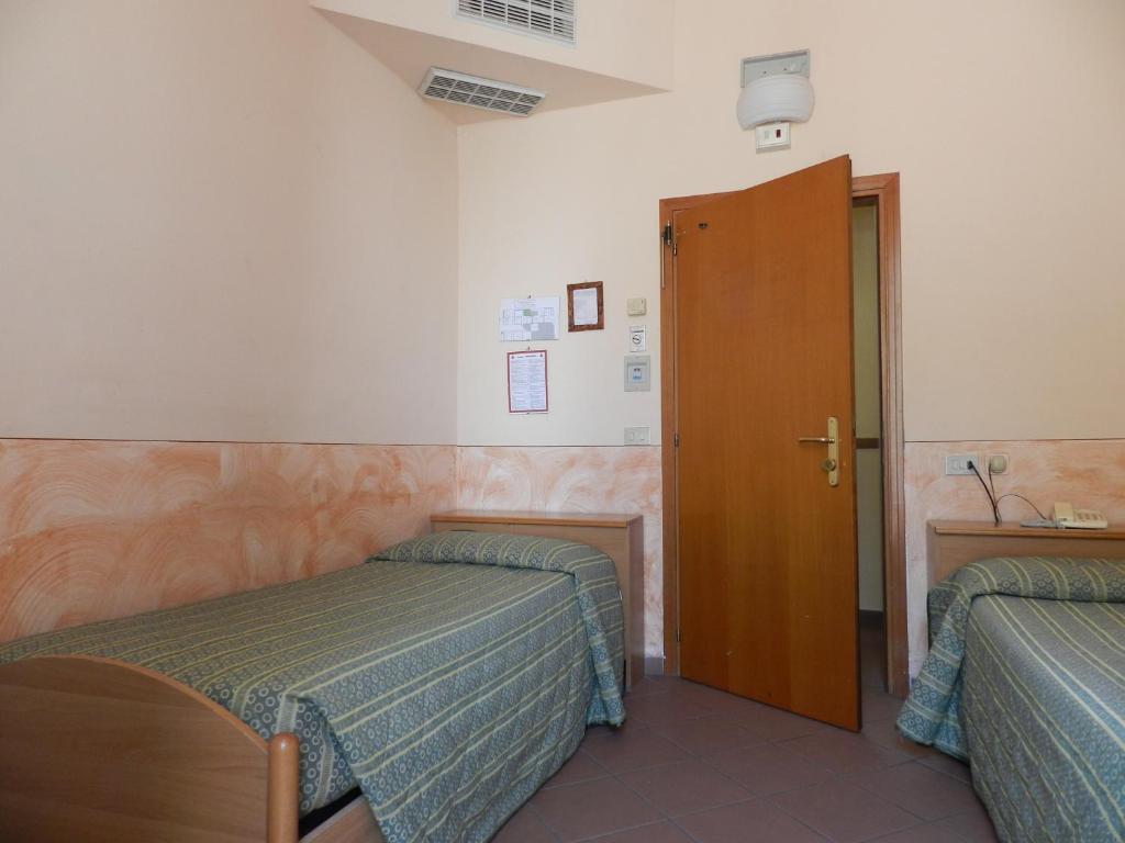 Divvietīgs numurs ar divām gultām, gaišām sienām un koka mēbelēm, kas nodrošina vienkāršu un ērtu nakšņošanu. Redzama atvērta durvju piekļuve citai telpai.