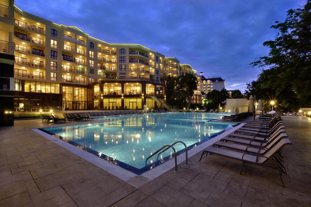 Moderns viesnīcas komplekss ar apgaismotām ēkām un peldbaseinu, kas atspoguļojas ūdenī. Vakarā redzams skats uz baseinu un atpūtas krēsliem.