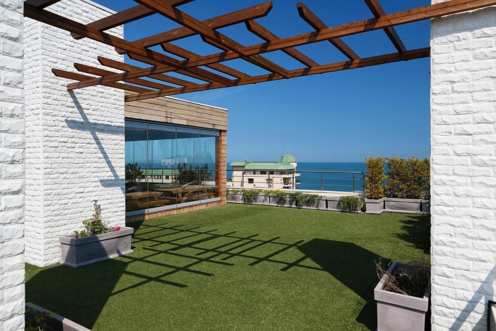 Mūsdienīgs jumta dārzs ar zāles segumu, dekoratīvām puķu podēm un koka pergolu, kas piedāvā skatu uz jūru. Ideāla vieta relaksācijai saulainā dienā.