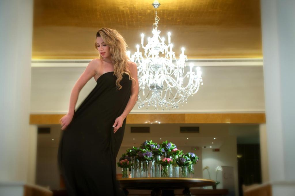Eleganta sieviete melnā kleitā pozē modernā interjerā ar lustru un ziedu kompozīciju fonā.