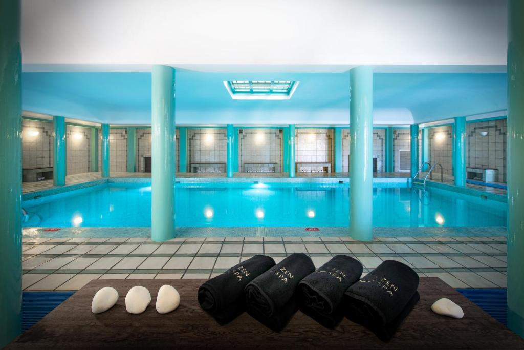 Mūsdienīgs iekštelpu peldbaseins ar skaidru ūdeni un stilīgām turquoise krāsas kolonnām. Blakus peldbaseinam ir salocīti dvieļi un akmeņi, kas rada relaksējošu atmosfēru.
