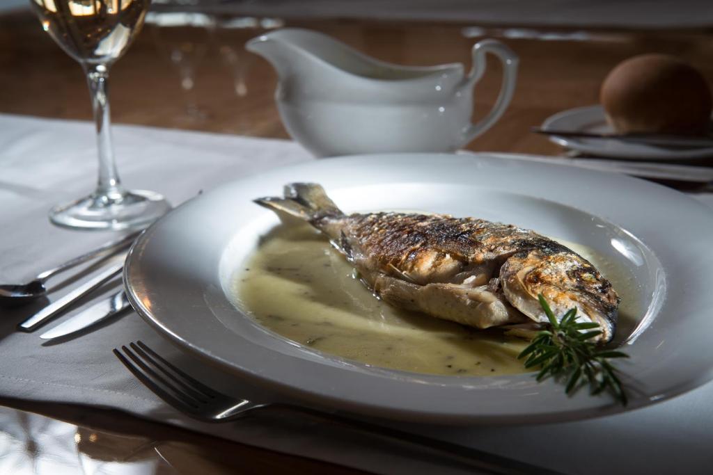 Grilēta zivs ar garšvielu mērci uz elegantas galda iestatījuma, papildināta ar vīna glāzi un maizes roll.
