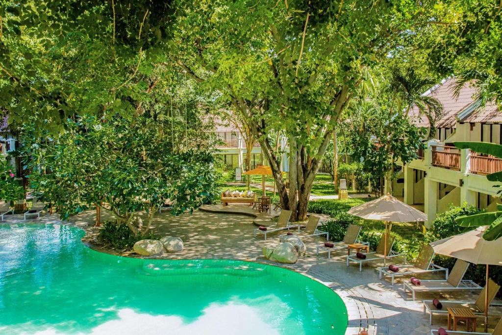 Tropu kūrorta ainava ar peldbaseinu, ko ieskauj zaļi koki un dārzi, radot mierīgu un relaksējošu atmosfēru. Gaisotne ir ideāla atpūtai un dabas baudīšanai.