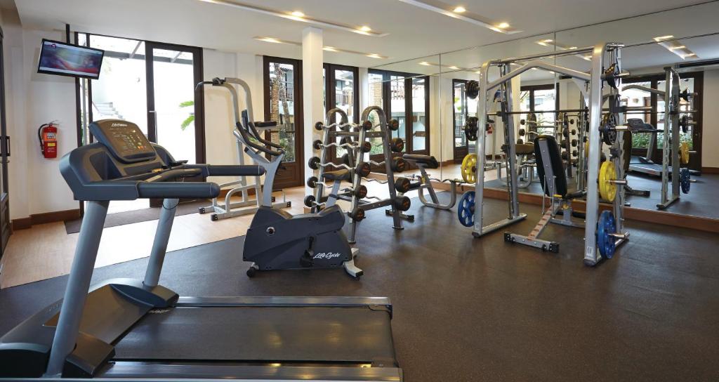 Mūsdienīgs sporta zāles interjers ar dažādu trenažieru aprīkojumu, tostarp skrejceliņu un svaru staciju. Telpa ir gaiša un plaša, ar logiem, kas nodrošina dabisko apgaismojumu.