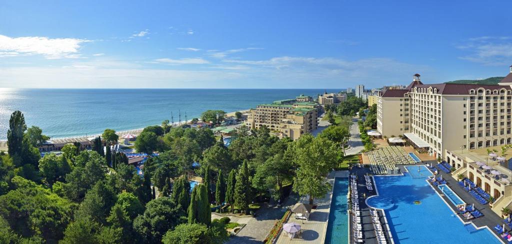 Jūras krastā redzama viesnīca ar plašu baseinu un zaļām ainavām, kas sniedzas līdz pludmalei. Skats uz saulainu debesi un mierīgu jūru.