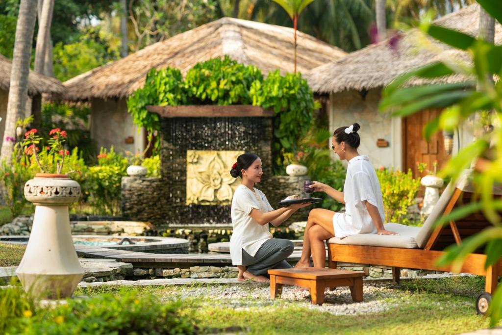 Divas sievietes relaksējas tropiskā dārzā, kur viena no tām pasniedz dzērienu. Fons ir skaista ainava ar ūdens elementiem un tradicionālām mājām.