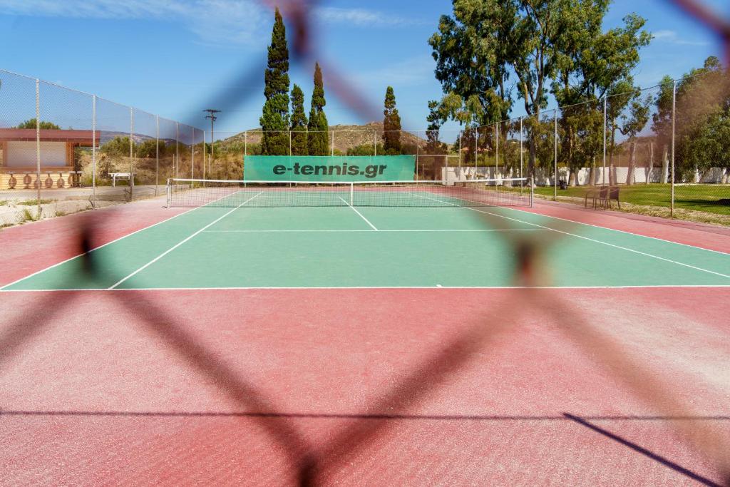 Tennis laukums ar zaļo segumu un sarkanu celiņu, redzams caur žoga acīm. Fons ir dabisks ar kokiem un kalniem, un laukuma vidū ir reklāma "e-tennis.gr".