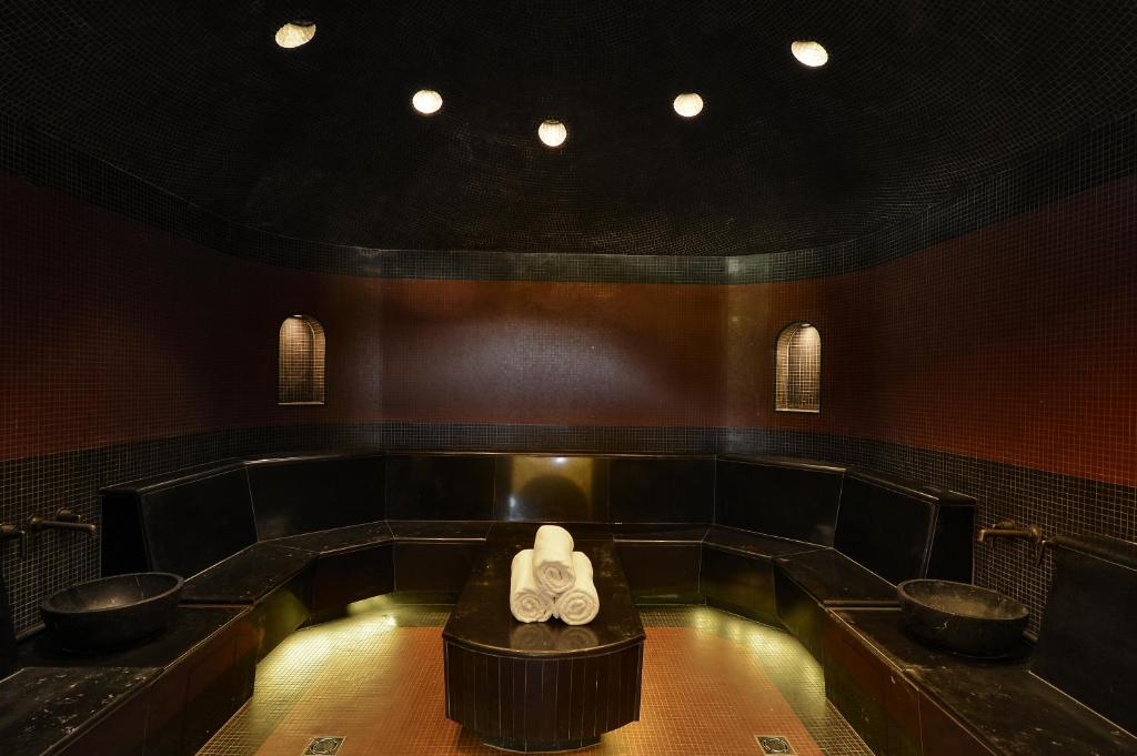 Mūsdienīga tvaika pirts ar melnajām flīzēm un sēdvietām, radot relaksējošu atmosfēru. Centrā novietoti dvieļi un divas melnas izlietnes.