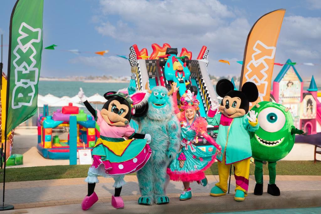 Priecīgas maskotās varoņi, tostarp Minnie un Mickey Mouse, kopā ar krāsainu dejotāju un citiem populāriem rakstzīmēm, svin jautrību tematiskajā parkā. Fons ir piepildīts ar spēļu iekārtām un karstām saule.