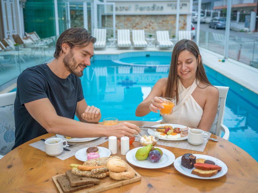 Pāris bauda brokastis pie galda pie peldbaseina, ar dažādiem ēdieniem un dzērieniem, tostarp augļiem un saldumiem. Gaisīgā atmosfērā ir siltas saules gaismas.